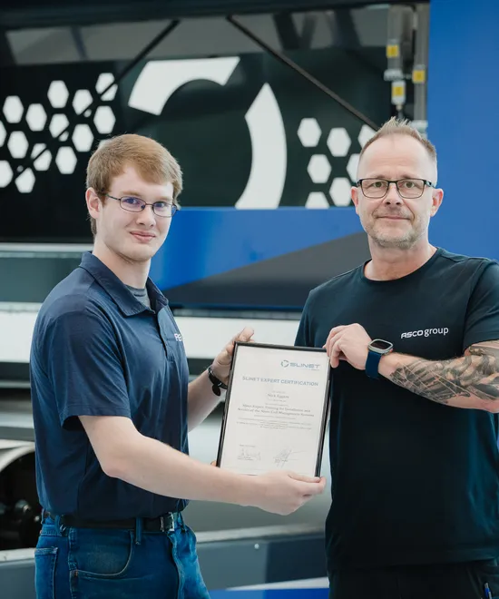 Zwei Mitarbeiter der ASCO Machinery Group mit Zertifikat vor einer Blechbearbeitungsmaschine in der Produktionshalle.